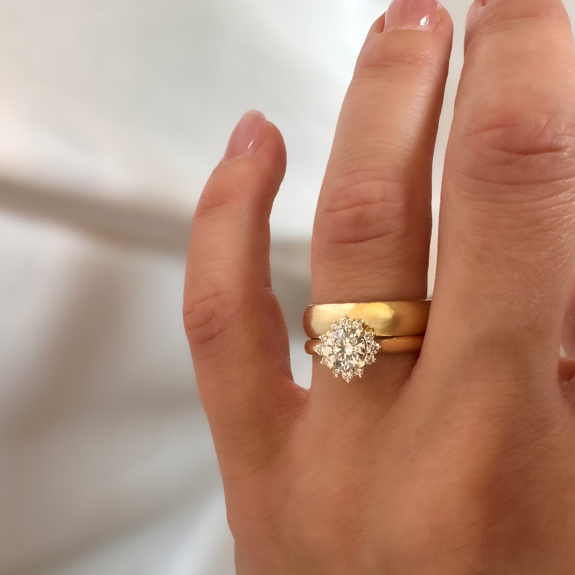 A person's hand displaying a floral engagement ring with a central diamond and smaller surrounding diamonds set in a 14K gold band stacked with gold wedding band.
