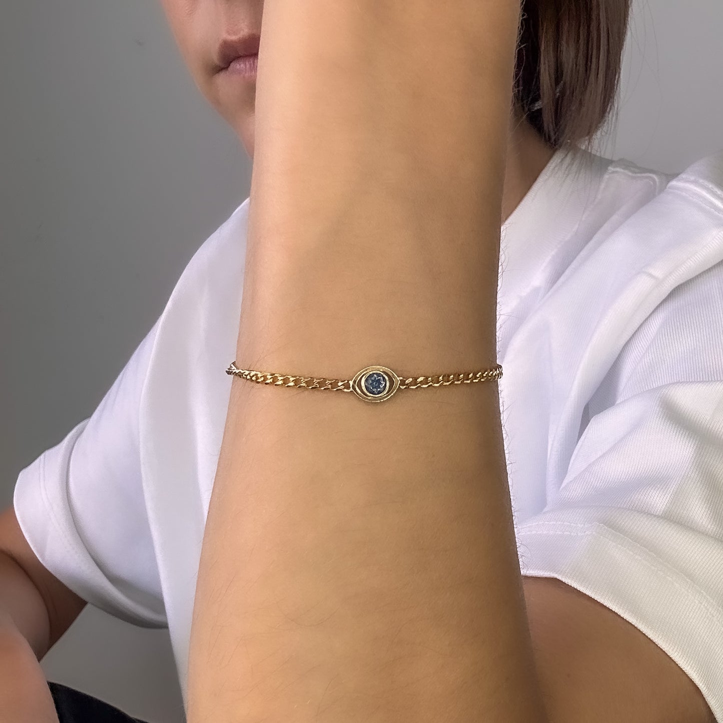 A person wearing a gold curb chain bracelet with a blue sapphire set in a evil eye centerpiece on their wrist.