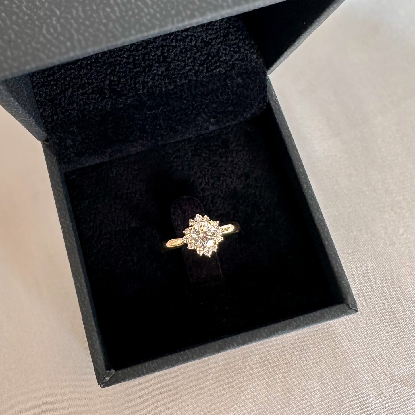 A floral engagement ring with a central diamond and smaller surrounding diamonds set in a 14K gold band in a black velvet box.