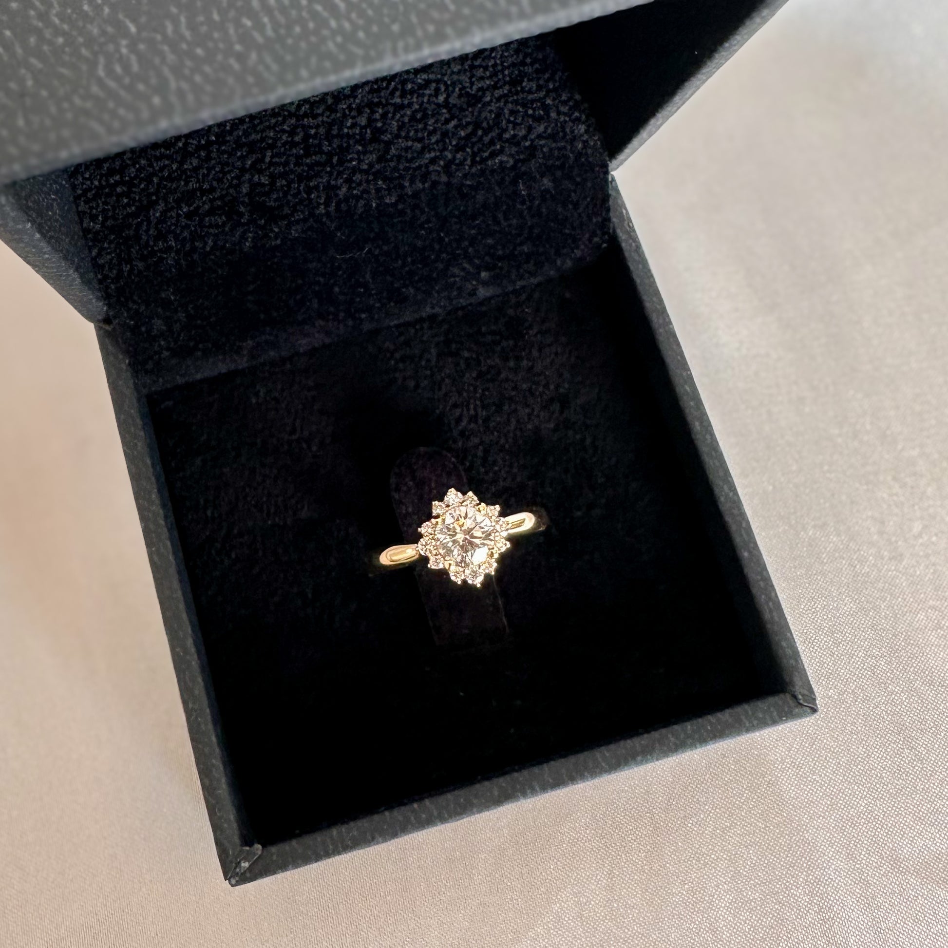 A floral engagement ring with a central diamond and smaller surrounding diamonds set in a 14K gold band in a black velvet box.