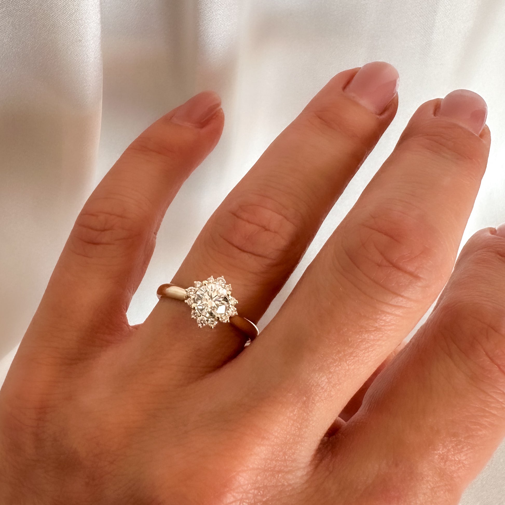 A person's hand displaying a floral engagement ring with a central diamond and smaller surrounding diamonds set in a 14K gold band.
