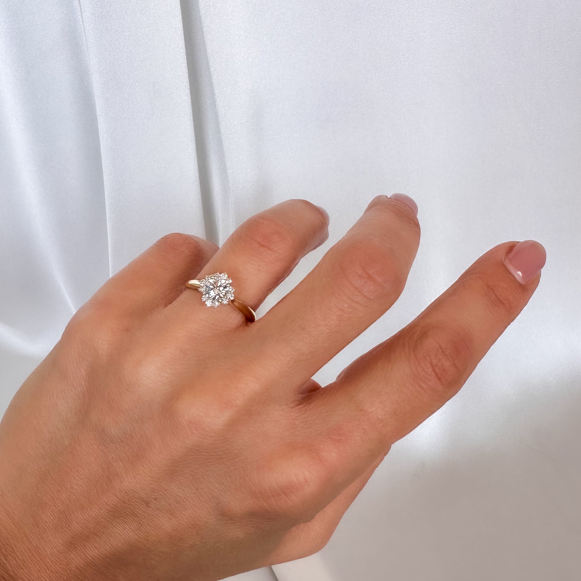 A person's hand displaying a floral engagement ring with a central diamond and smaller surrounding diamonds set in a 14K gold band.