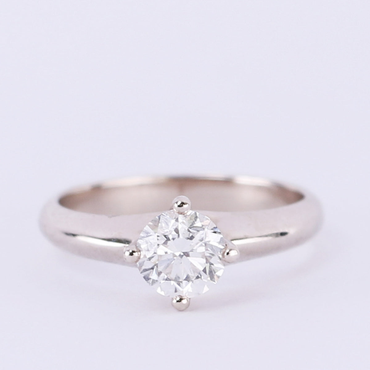 Four-prong round diamond solitaire engagement ring in white gold on a light purple background