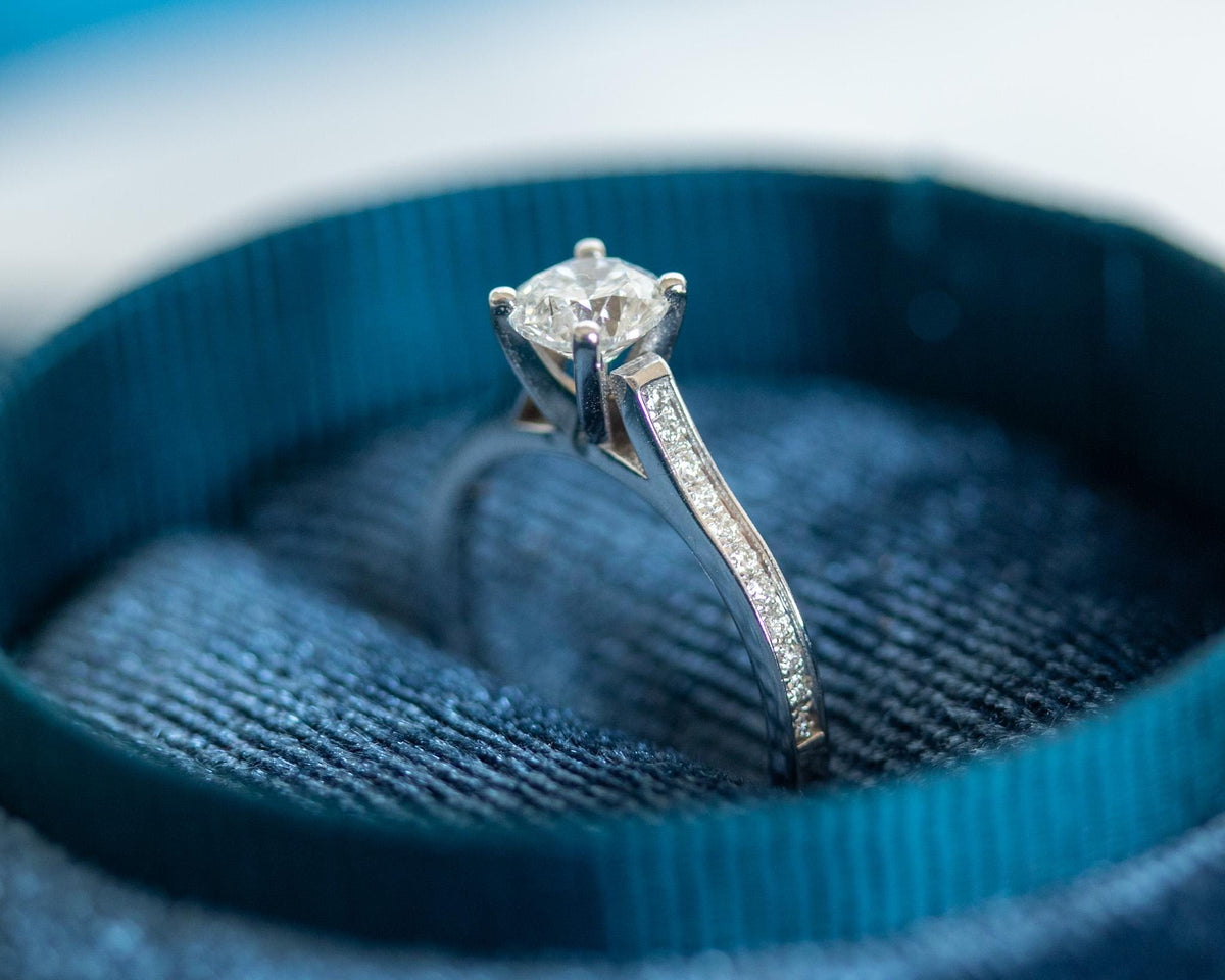 Four-prong round diamond ring with channel-set diamonds on the band, white gold, side view in a blue box