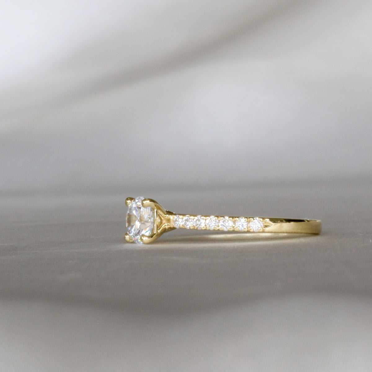 Side view of a four-prong round diamond ring with pavé diamonds on the band, yellow gold
