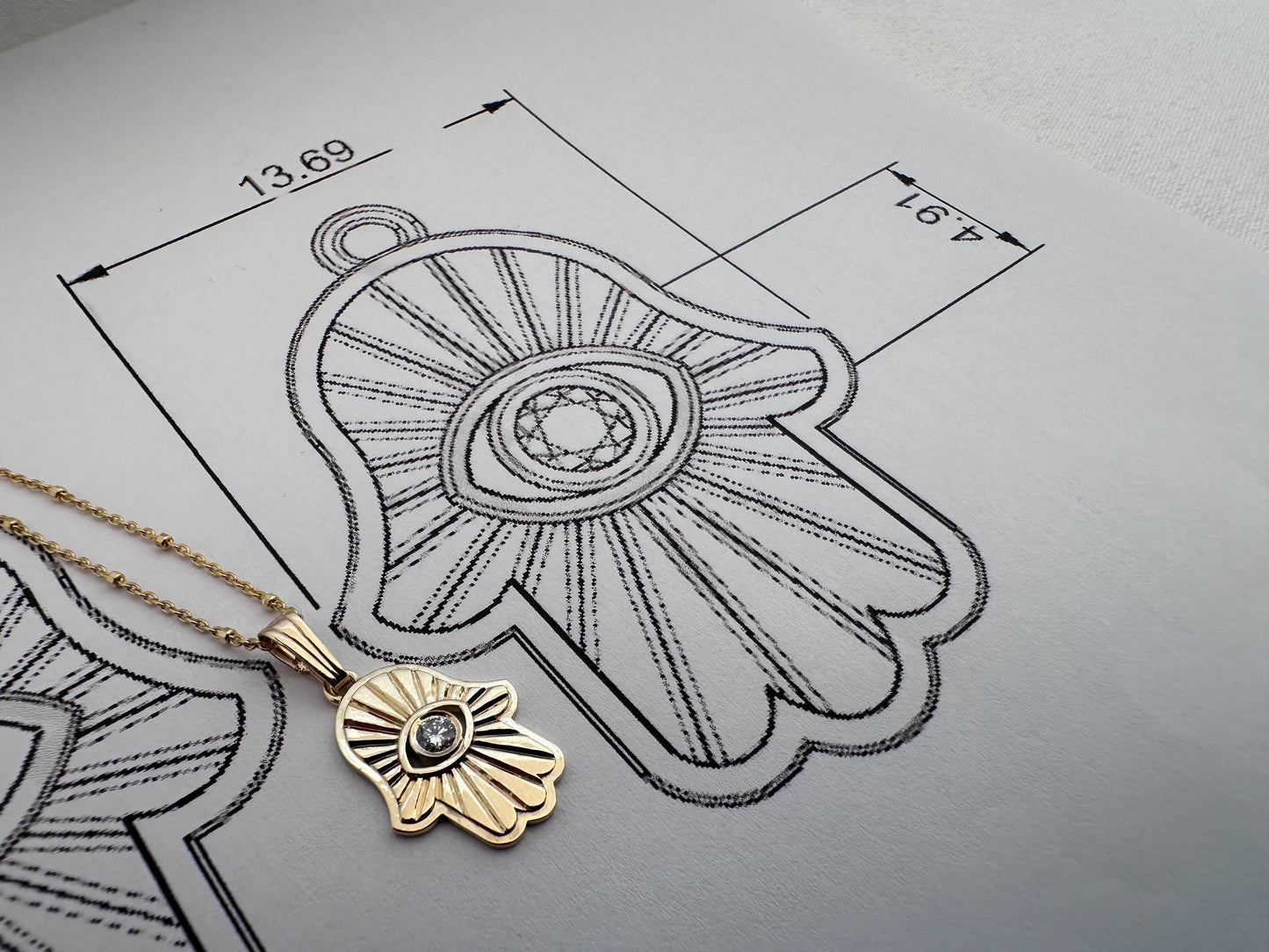 Gold pendant on a gray surface with technical drawing and measurements