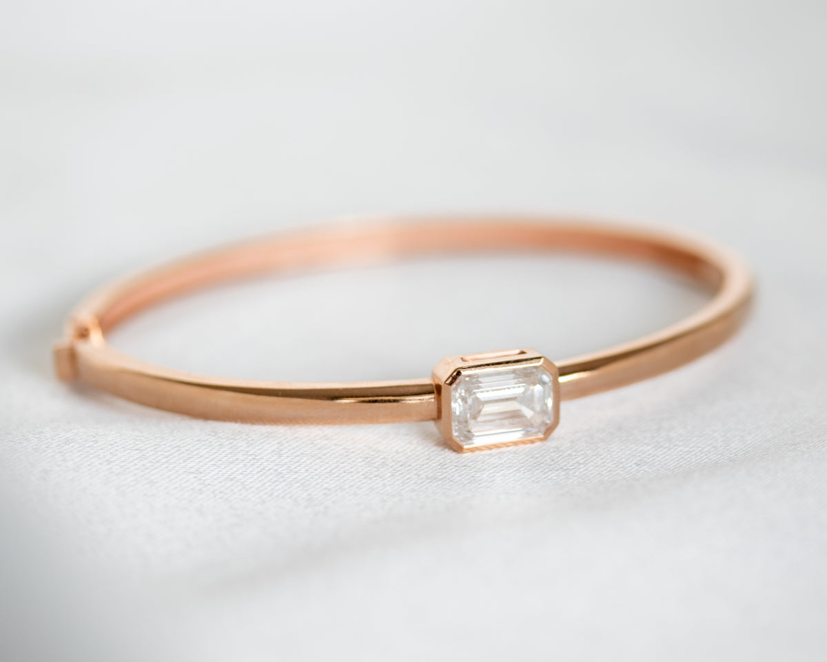 14K gold bezel-set emerald cut diamond bangle, diagonal view on light gray background