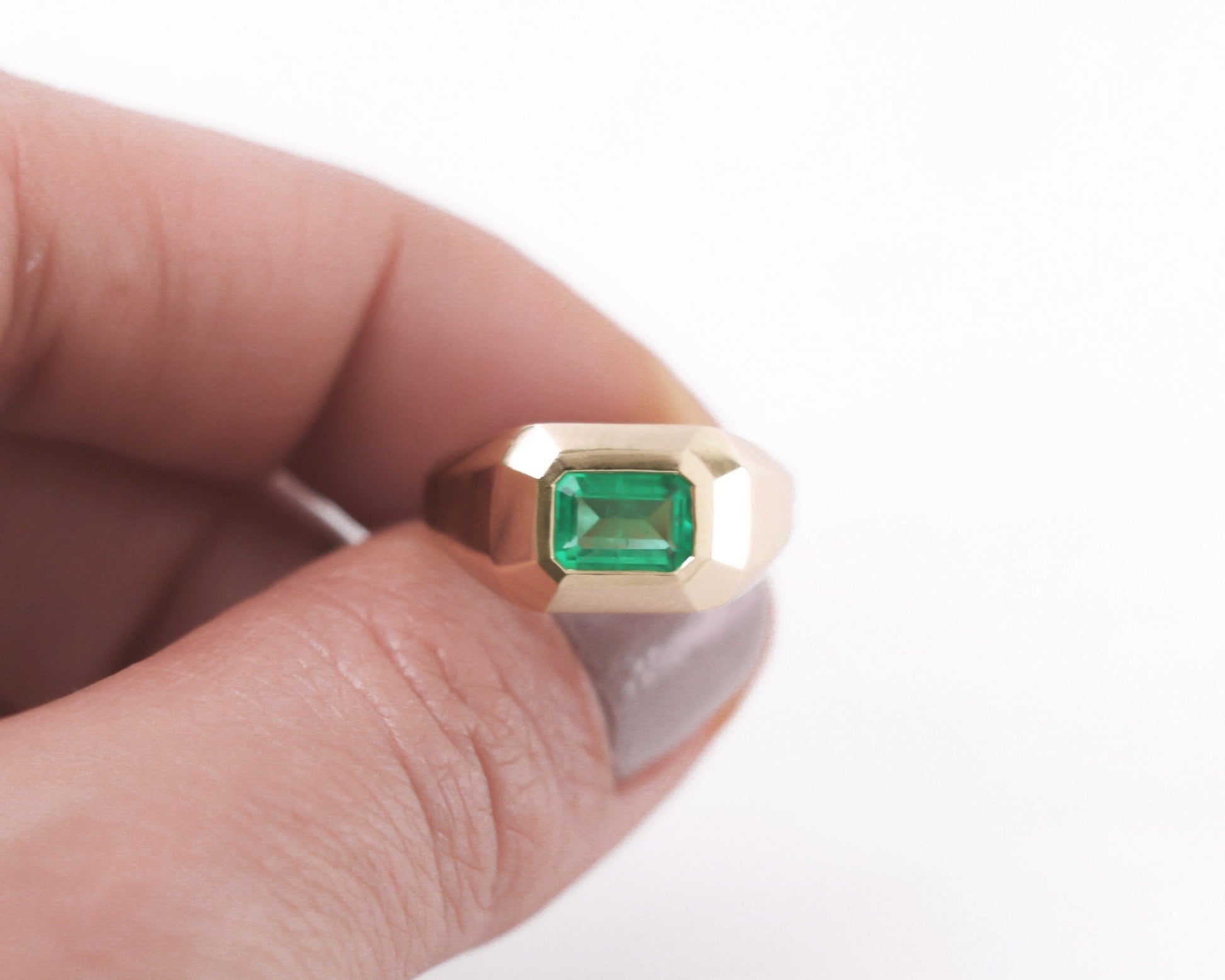 Gold men’s signet ring with emerald-cut green gemstone held between fingers against a white background