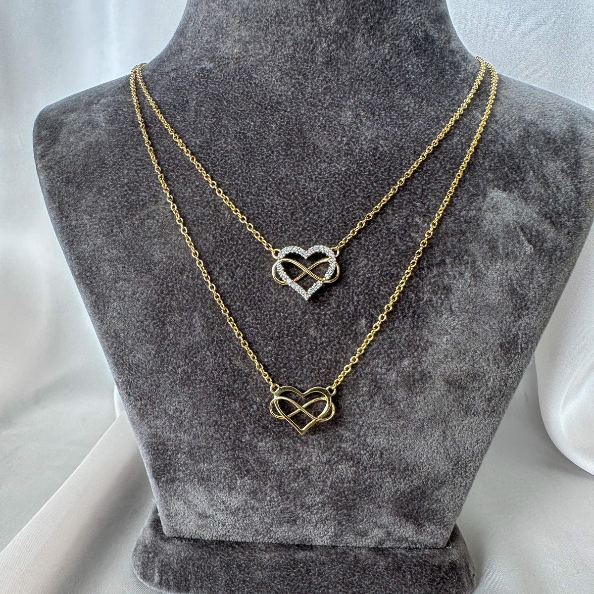 Two gold heart necklaces layered on a gray jewelry bust