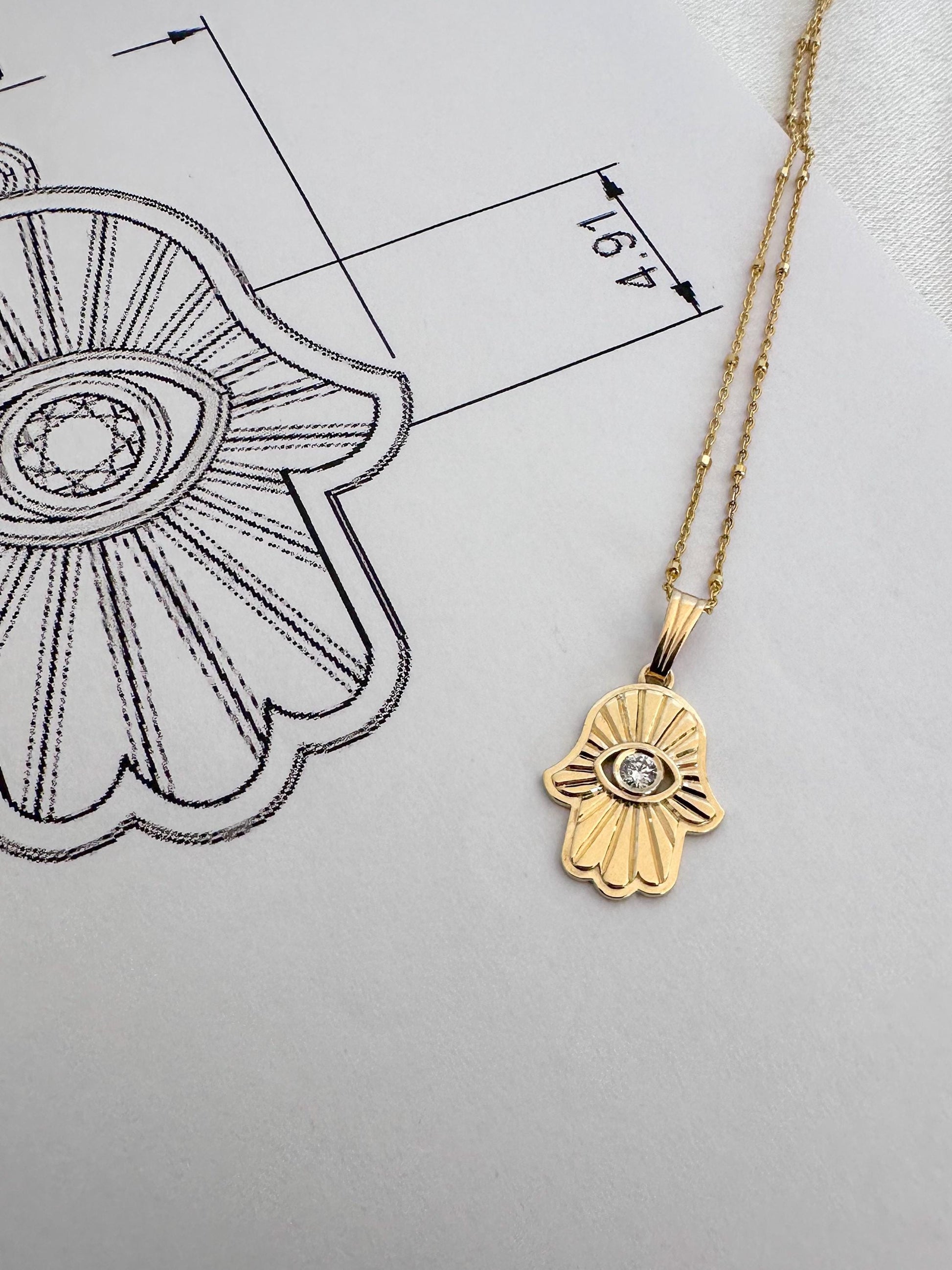 Gold Hamsa hand necklace with a diamond on a white background with a black line drawing of an eye.