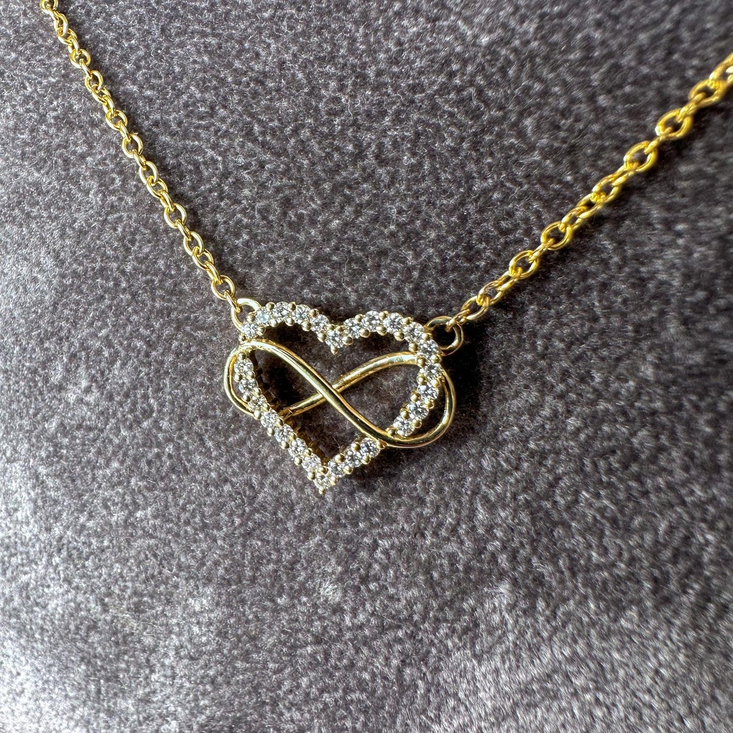 Gold necklace with a heart and infinity symbol pendant on a gray textured background
