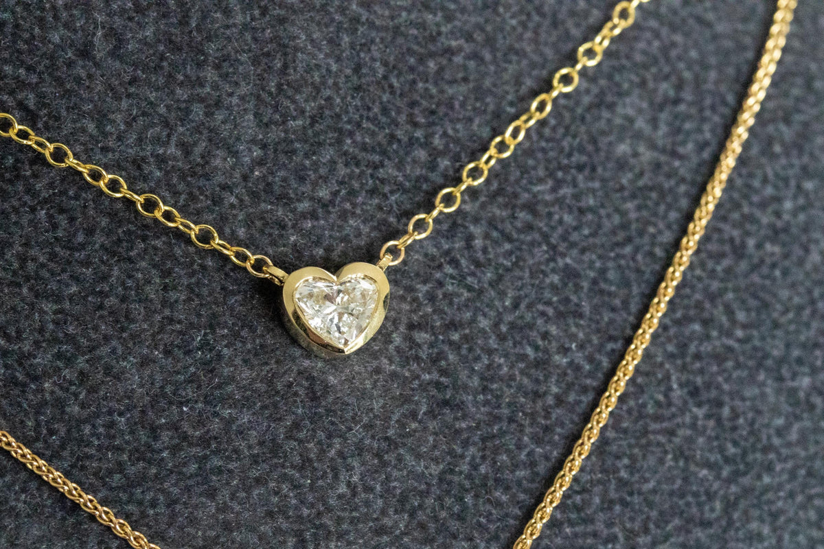 Heart shape diamond in a bezel setting on a 14K yellow gold rolo chain, layered with a second chain on gray velvet