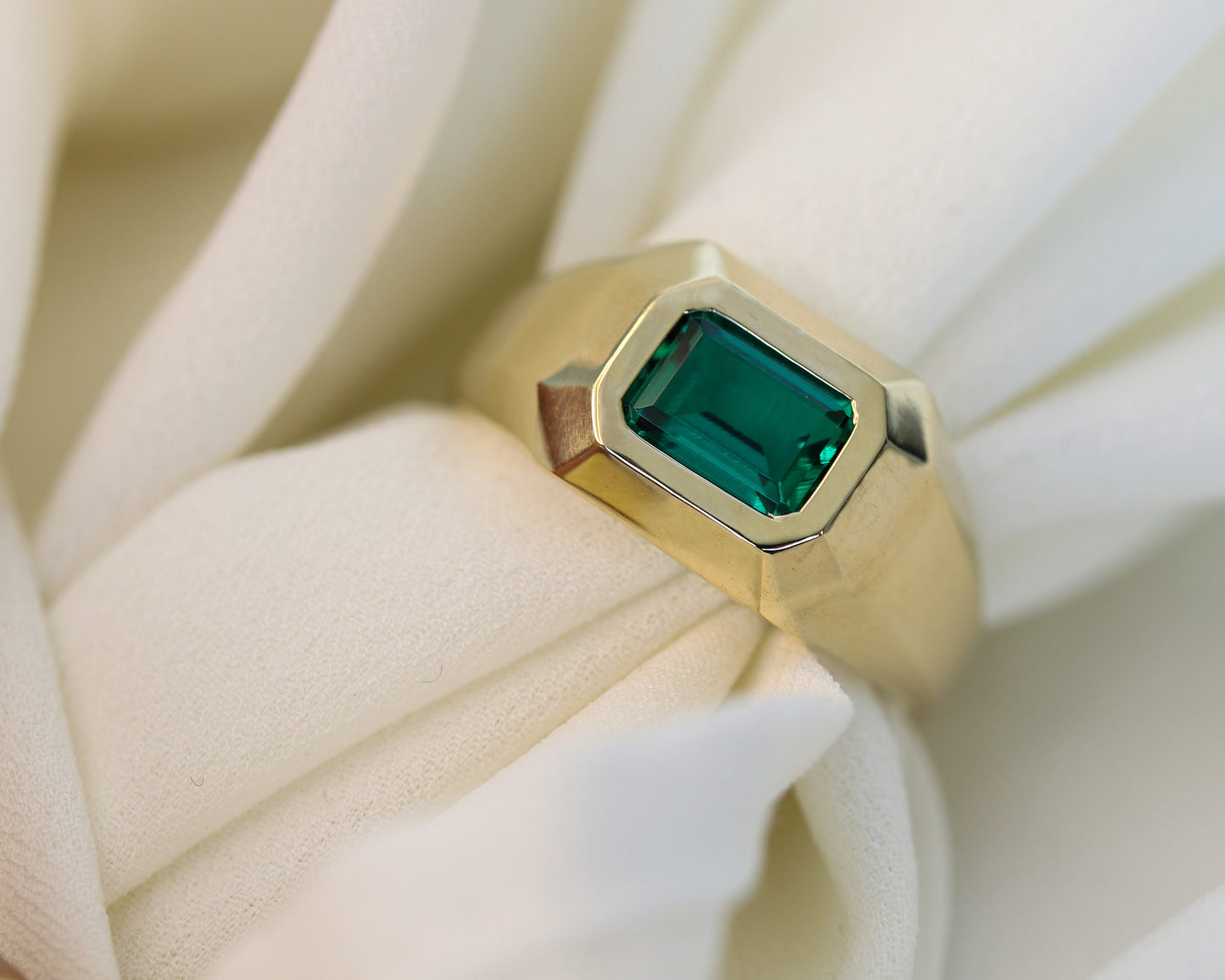 Men’s 14K gold signet ring with an emerald-cut green emerald on a soft white fabric background.
