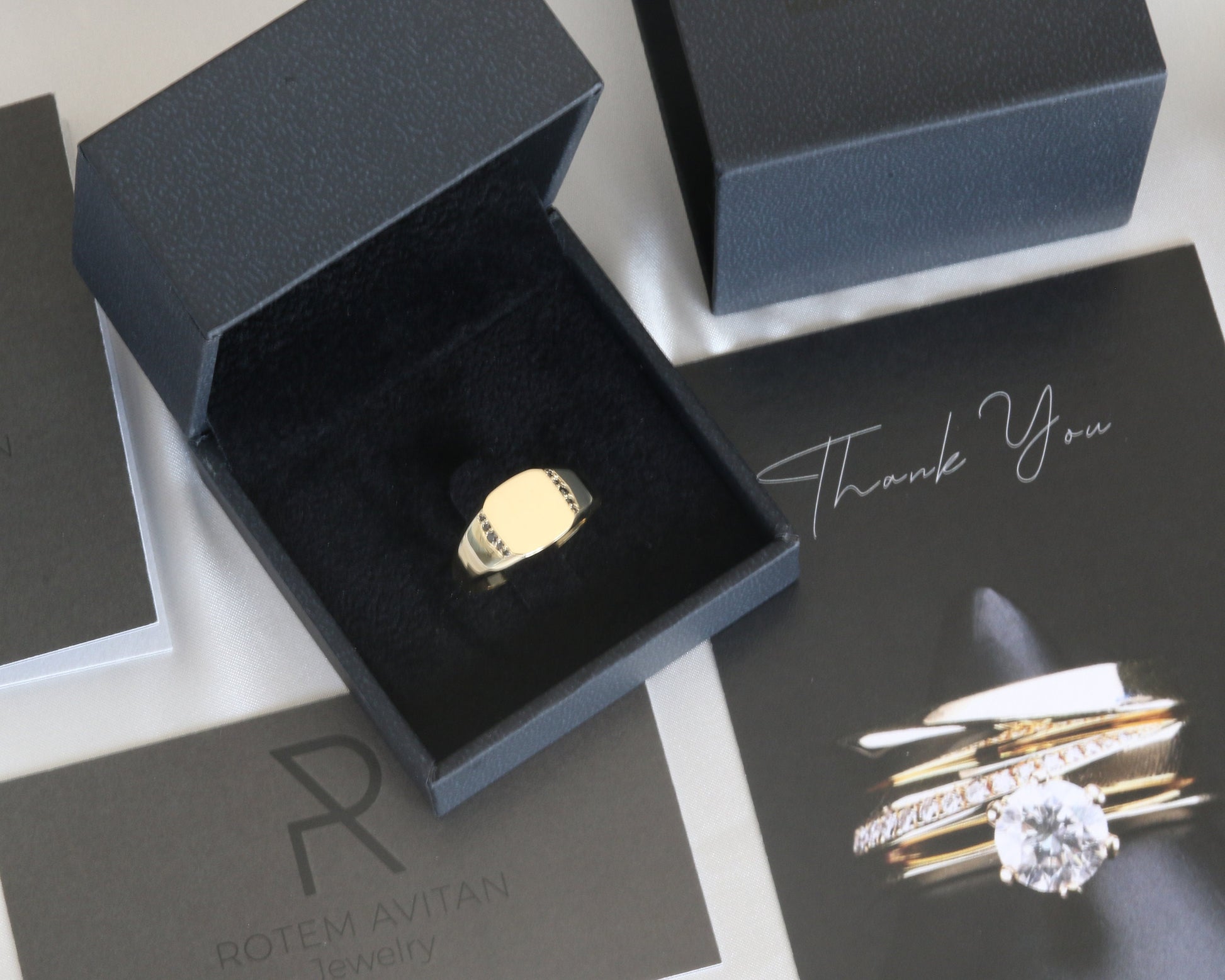 Men’s 14K gold signet ring with black diamonds set along the sides, displayed in a jewelry box with branding materials.