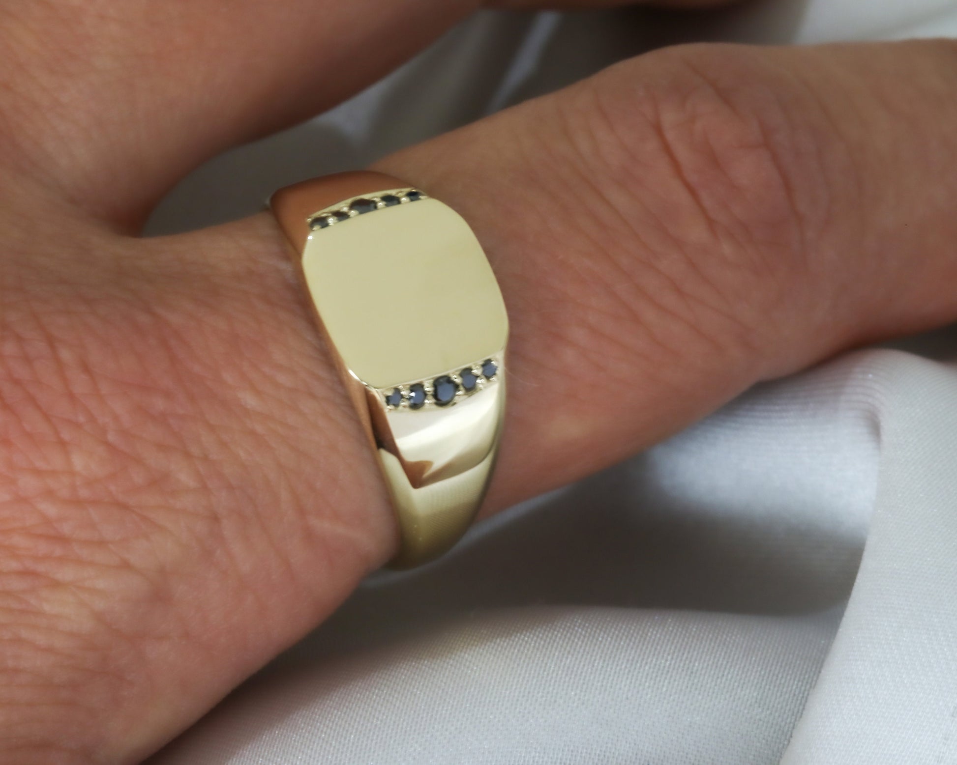 Men’s 14K gold signet ring with black diamonds set along the sides, worn on a hand against a soft white fabric background.