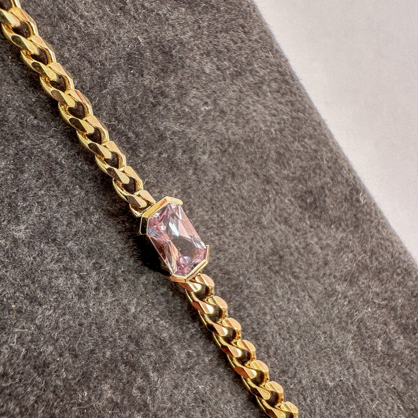 A gold bracelets with a curb chain design featuring an emerald cut natural pink sapphire displayed on a gray textured stand