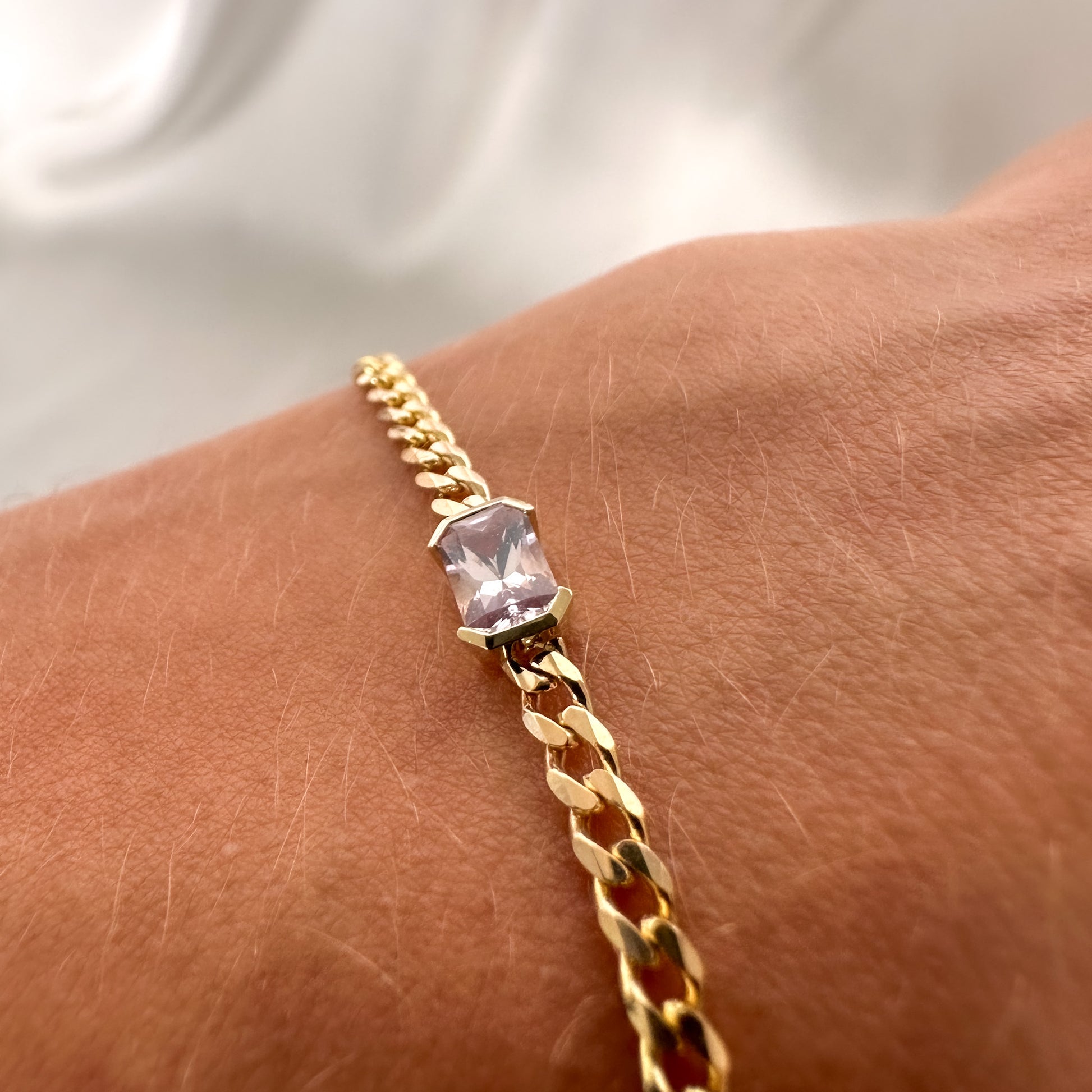A gold bracelets with a curb chain design one featuring an emerald cut natural pink sapphire displayed on a person's wrist.