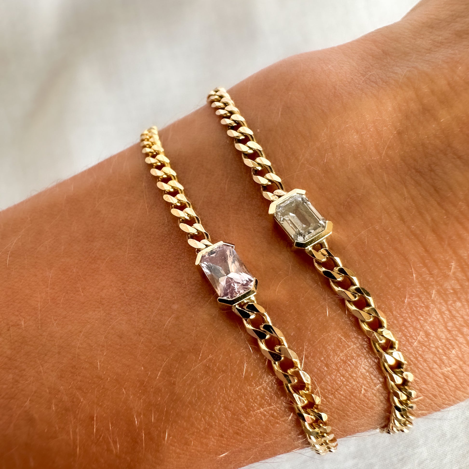 Two gold bracelets with a curb chain design one featuring an emerald cut diamond solitaire, and the other  featuring an emerald cut natural pink sapphire displayed on a person's wrist.