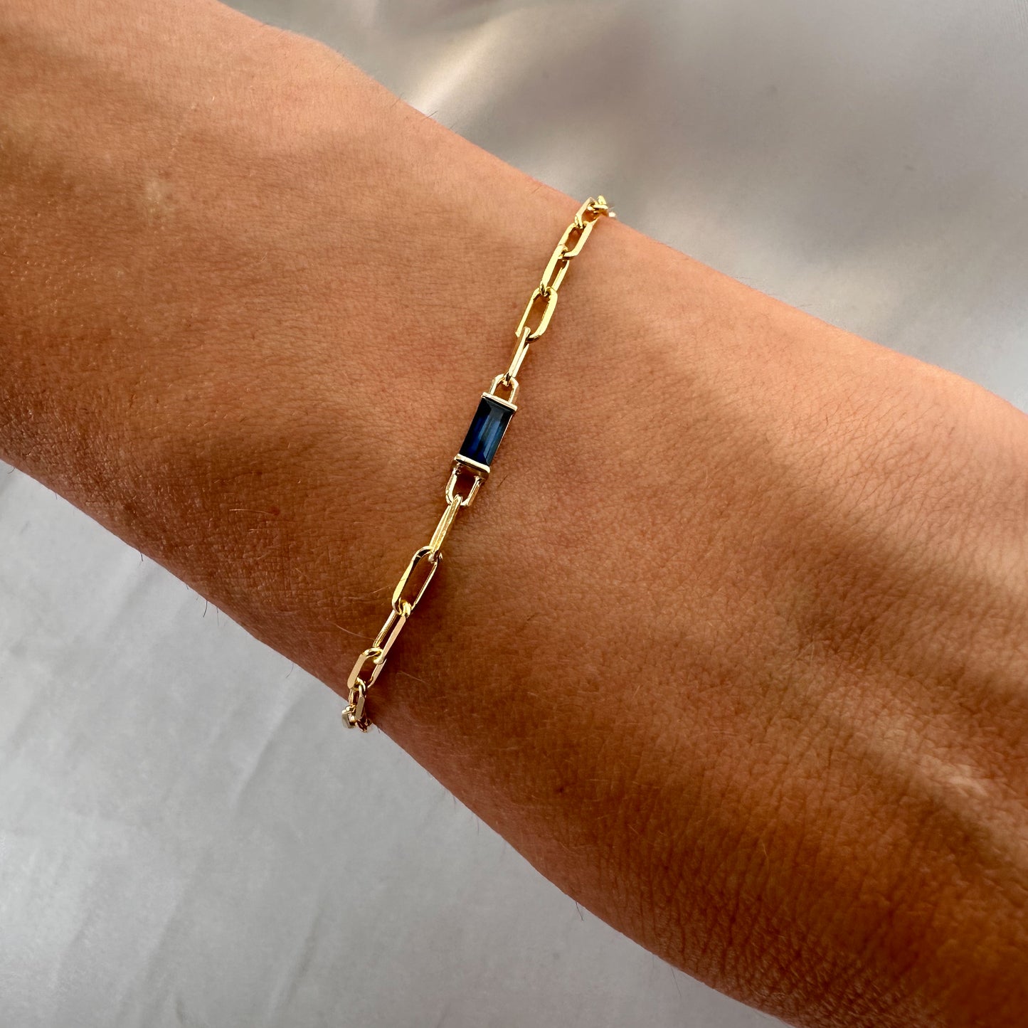 A gold bracelet featuring a baguette cut blue sapphire stone, with a chain of paper clips attached to it, worn on a wrist.