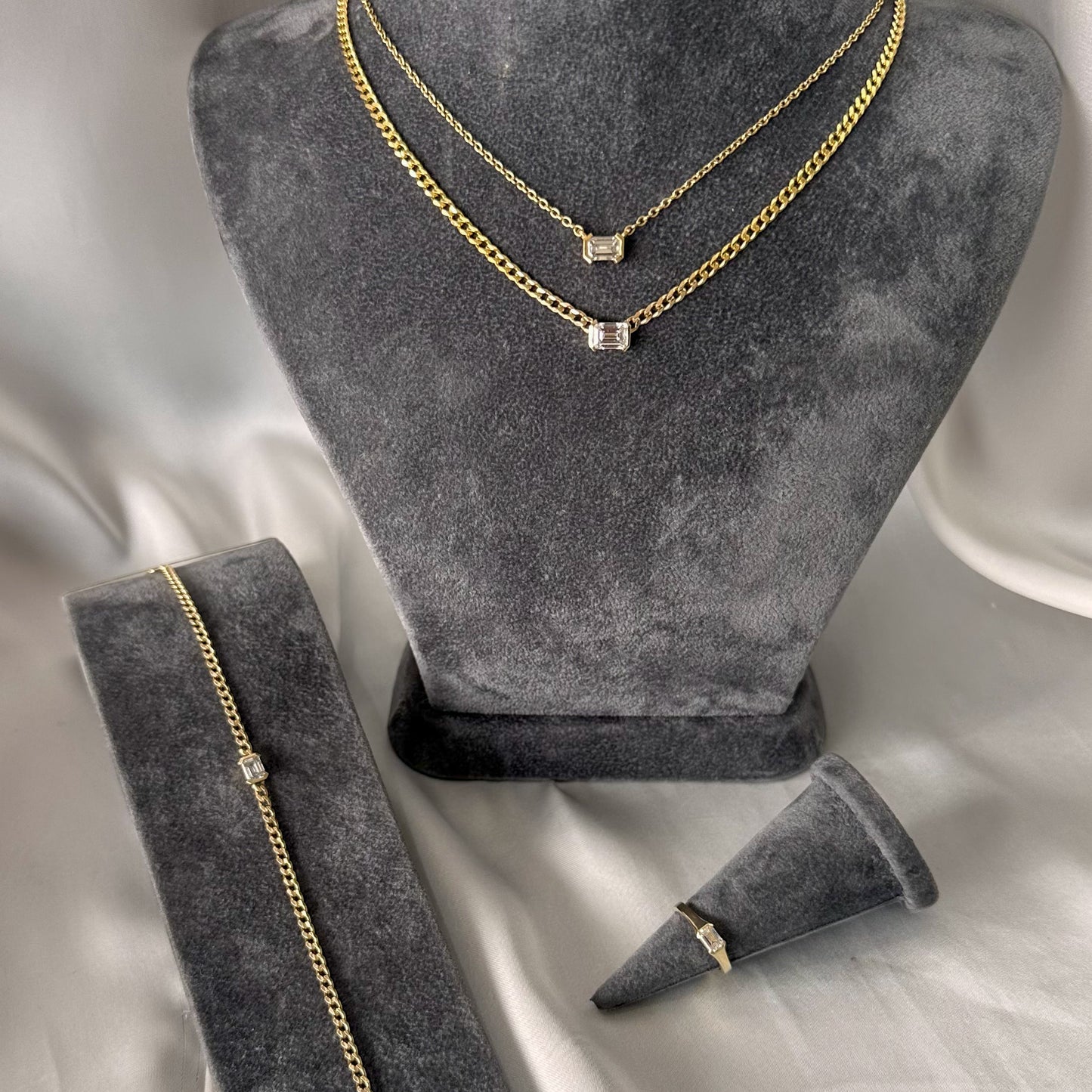 A 14K gold diamond solitaire necklace with an emerald cut natural diamond pendant on a gray textured jewelry torso emerald cut diamond. ring on gray textured stand and gold curb chain bracelet with emerald cut diamond on gray textured stand