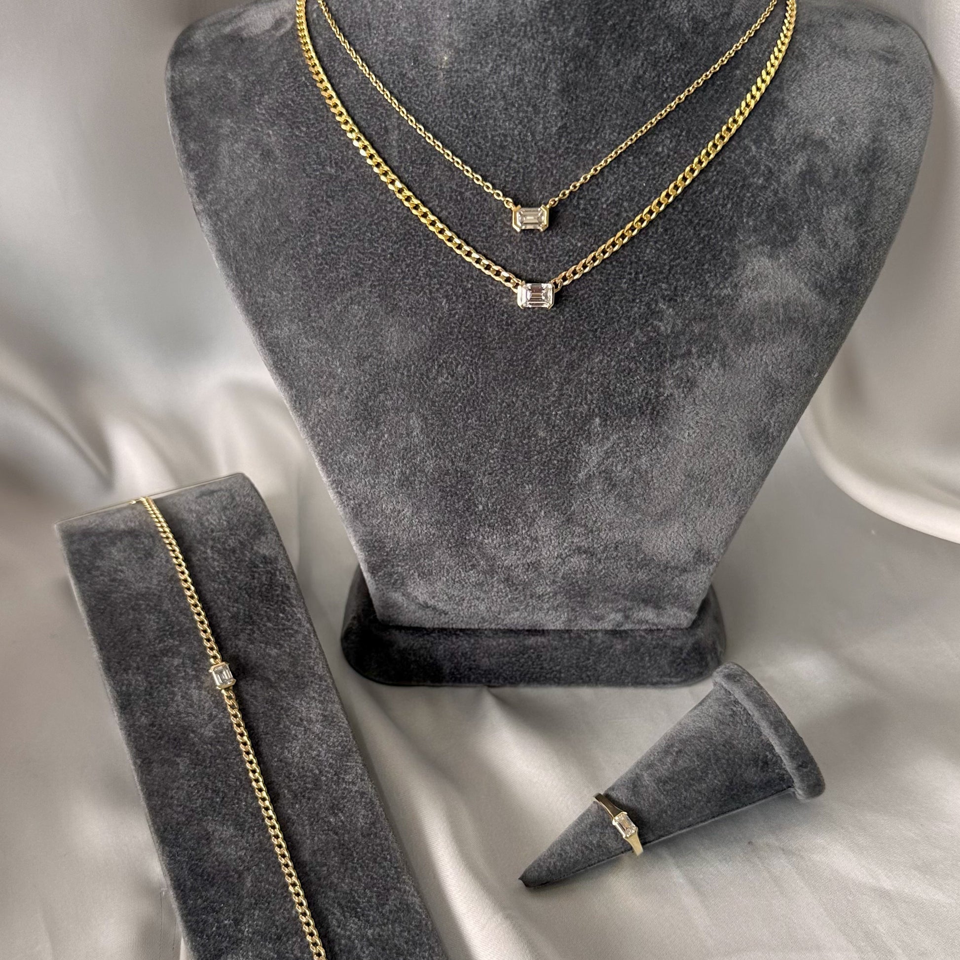 A 14K gold diamond solitaire necklace with an emerald cut natural diamond pendant on a gray textured jewelry torso emerald cut diamond. ring on gray textured stand and gold curb chain bracelet with emerald cut diamond on gray textured stand