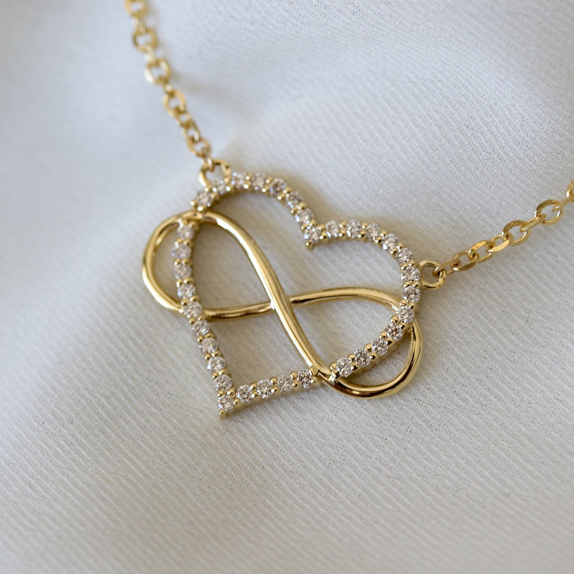 A heart-shaped pendant necklace with a diamonds and gold infinity symbol, attached to a yellow gold chain, displayed on white background