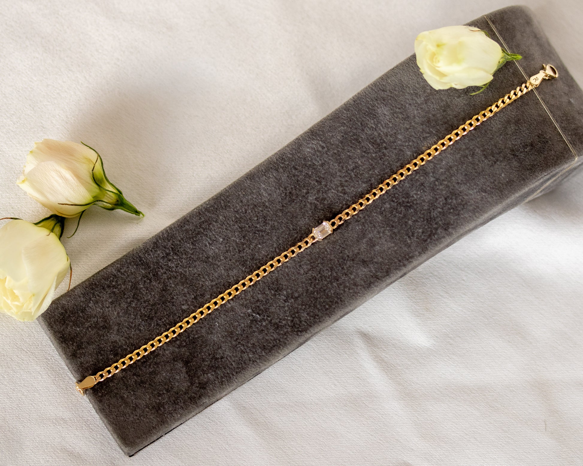 Gold bracelet with an emerald cut diamond on a gray surface with white roses