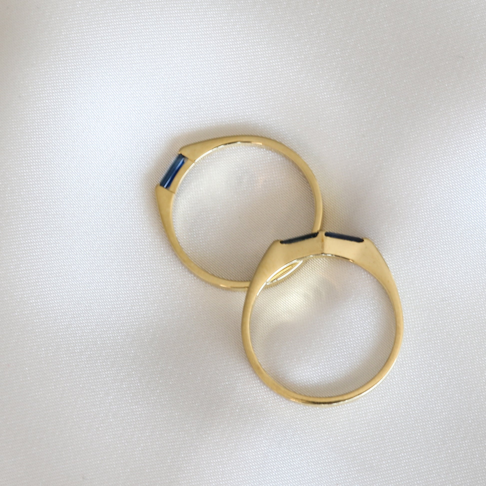 A minimalist style of two rings with needle baguette cuts blue sapphires set in a 14K yellow gold band, displayed against a white background.