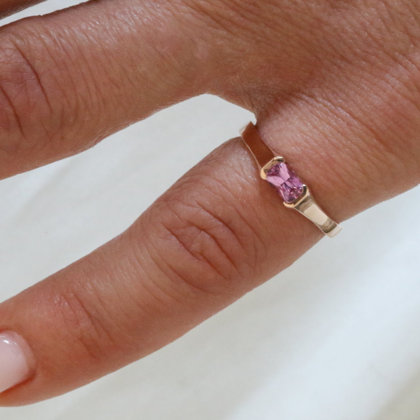 A rose gold ring with a emerald cut natural pink sapphire stone, set in a half bezel setting on women's pinky finger