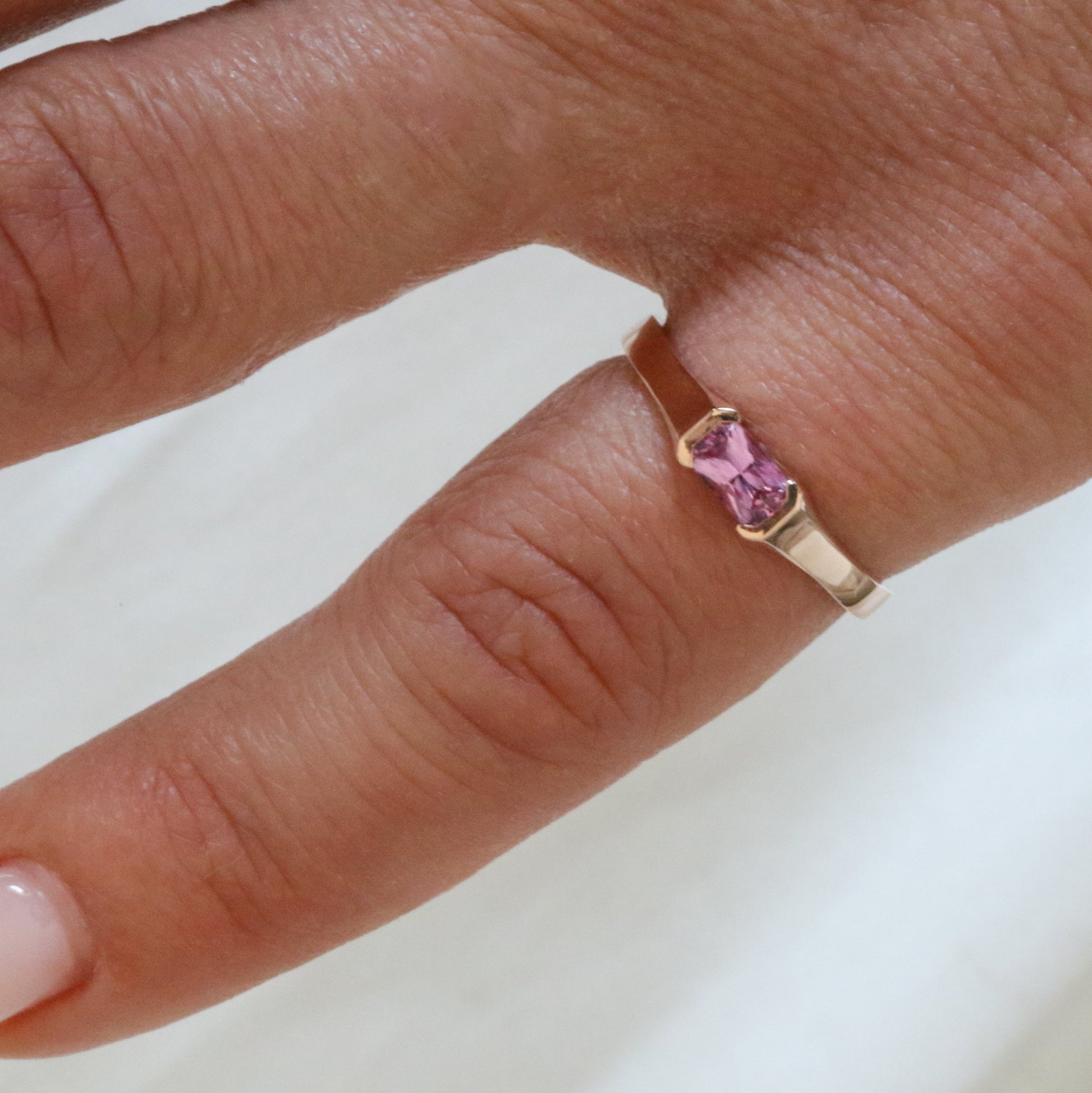 A rose gold ring with a emerald cut natural pink sapphire stone, set in a half bezel setting on women's pinky finger
