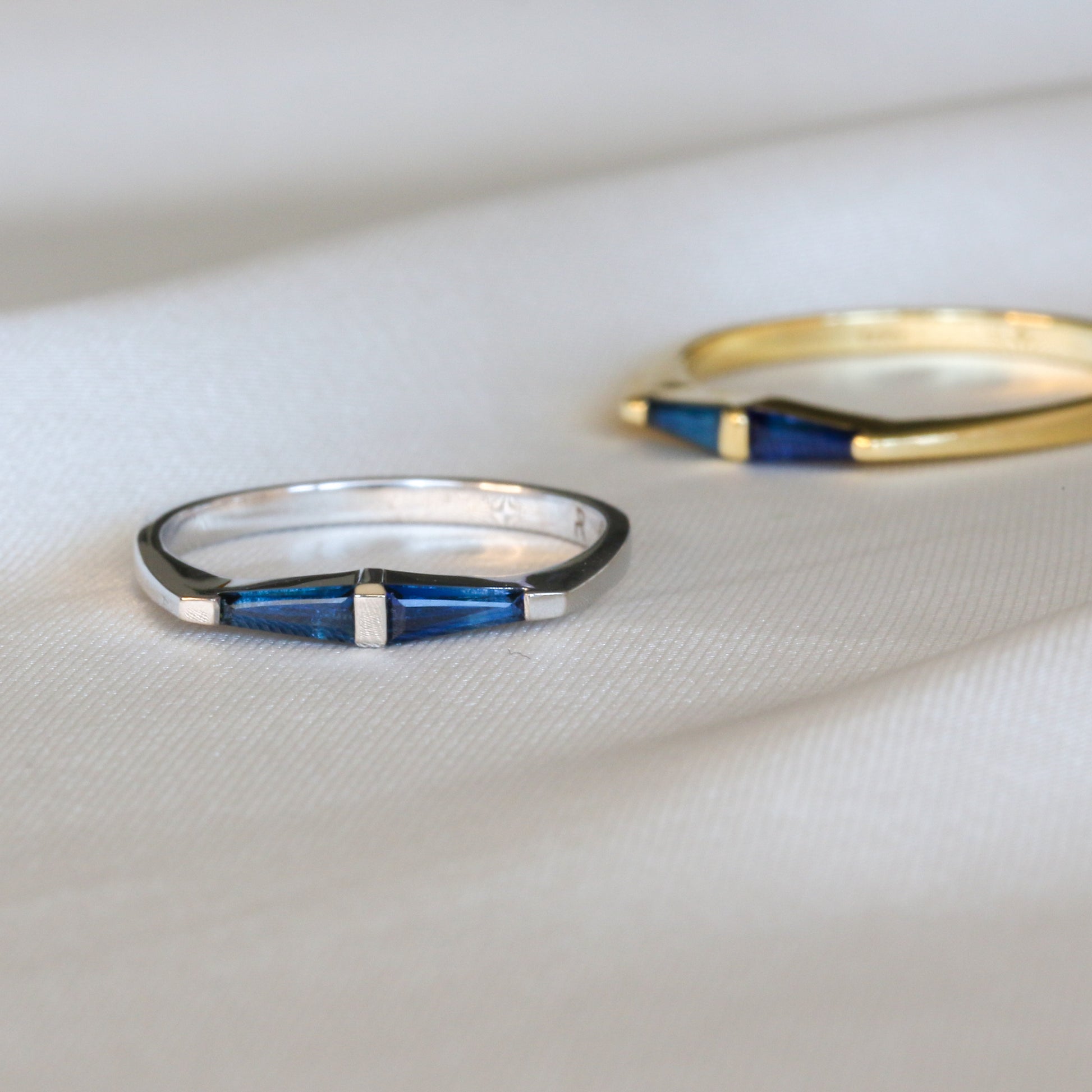 A 14K white gold ring with two natural blue tapered baguette cut sapphires with same ring in yellow gold in the background