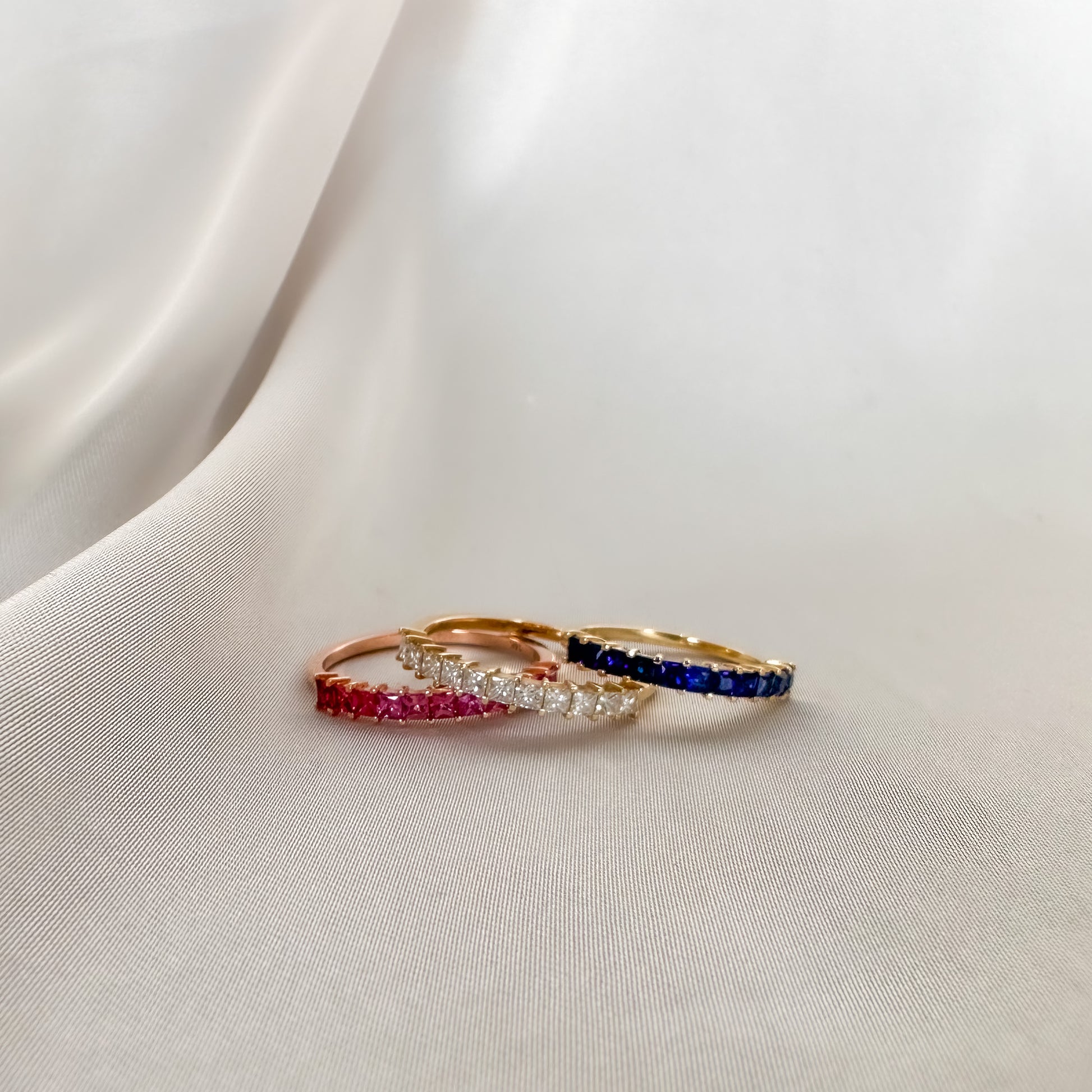 three princess cut eternity rings each set with different type f stone. diamond, pink sapphire and blue sapphire on white cloth
