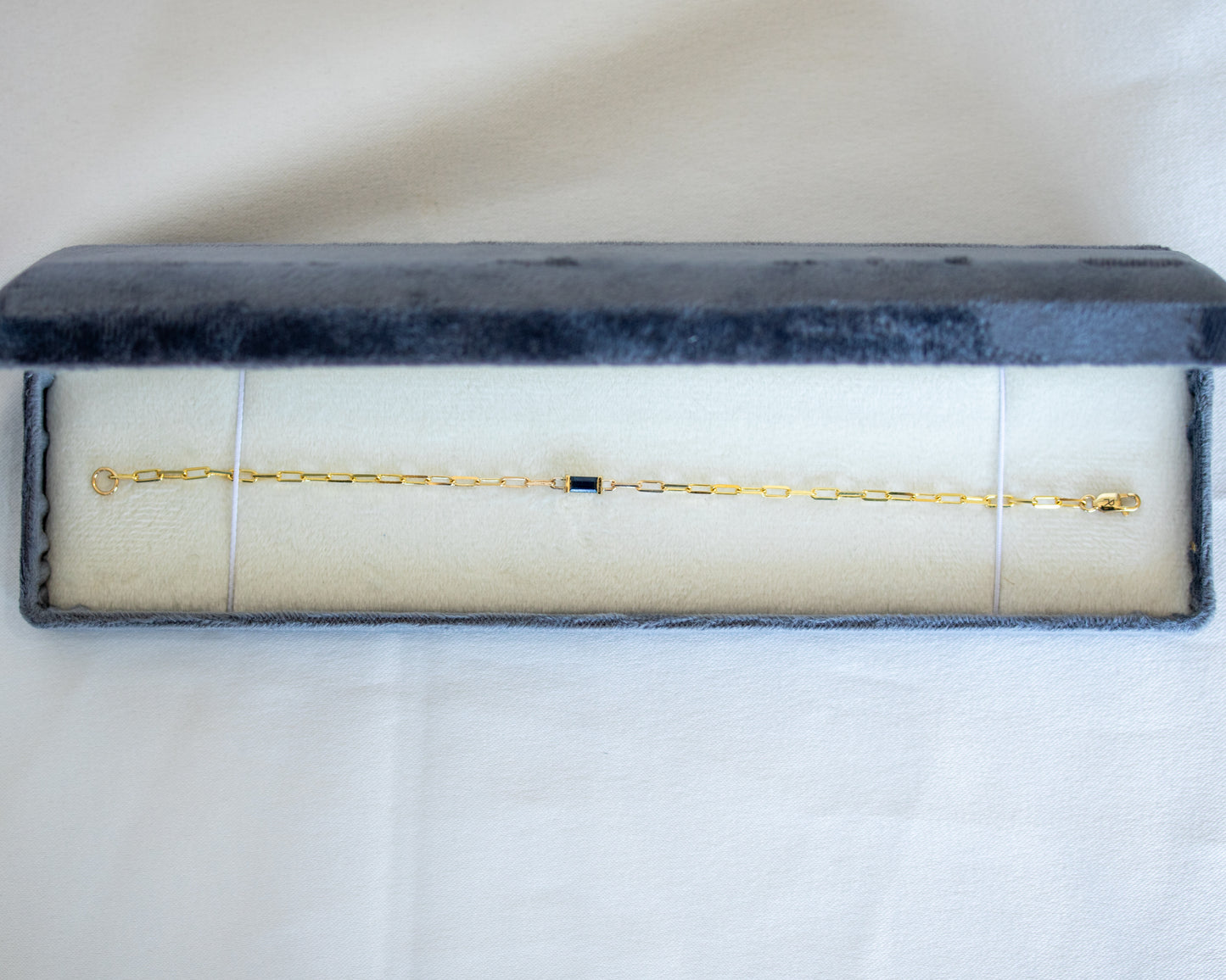 Gold bracelet with a blue baguette cut sapphire in a gray velvet box on a white background