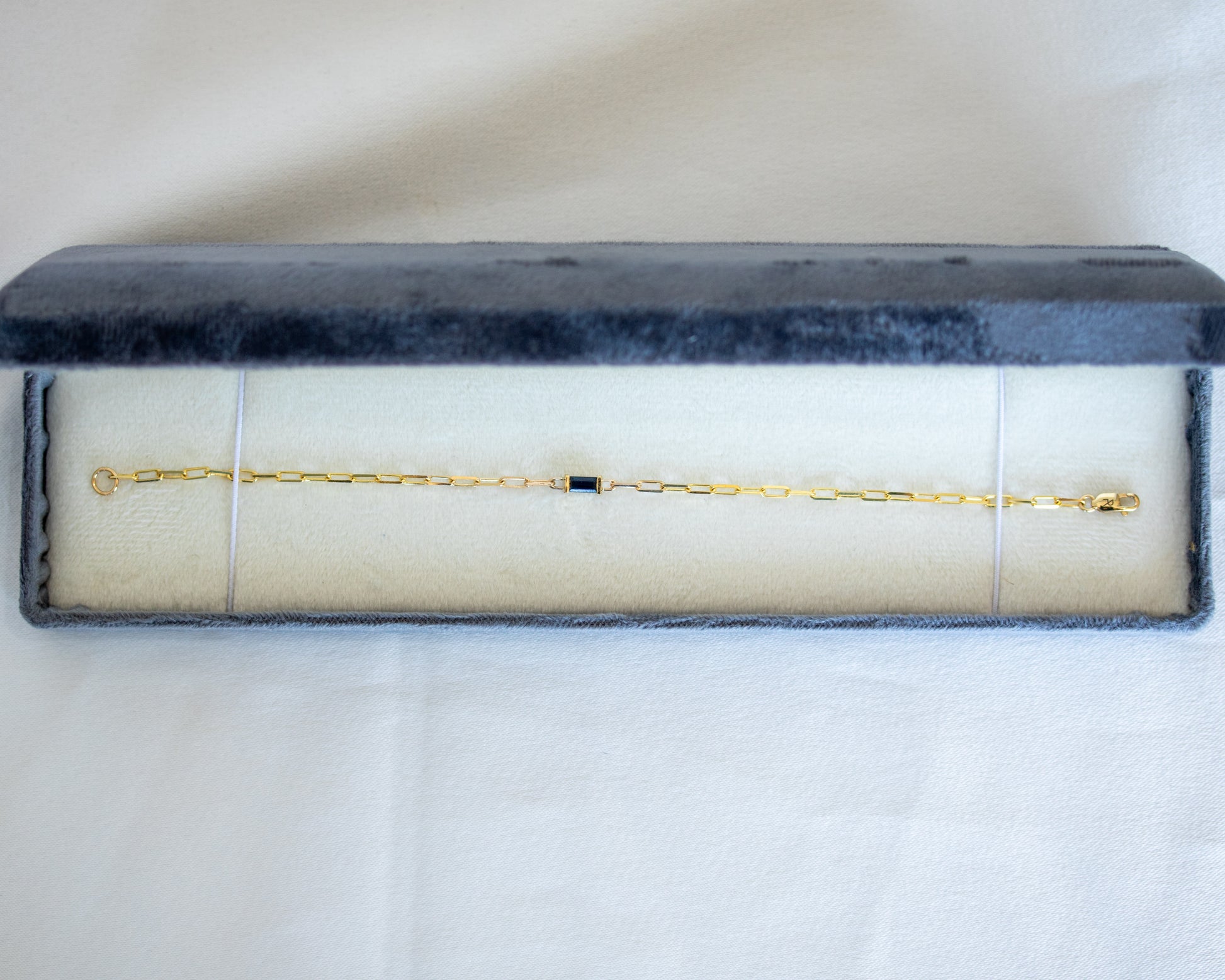 Gold bracelet with a blue baguette cut sapphire in a gray velvet box on a white background
