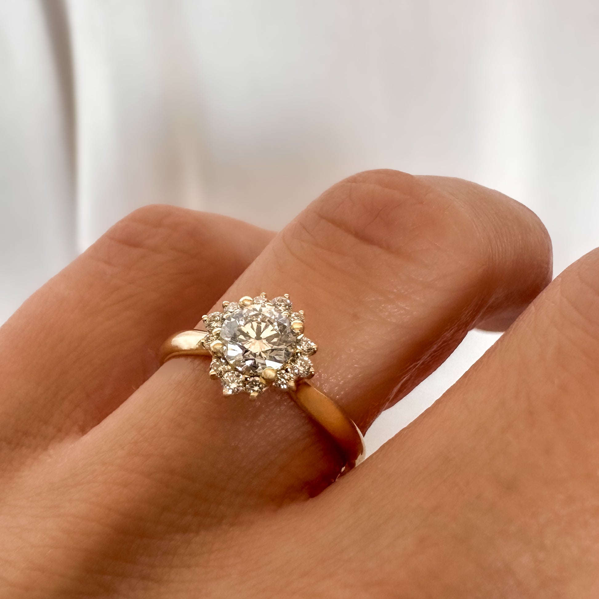close up of a person's hand displaying a floral engagement ring with a central diamond and smaller surrounding diamonds set in a 14K gold band.