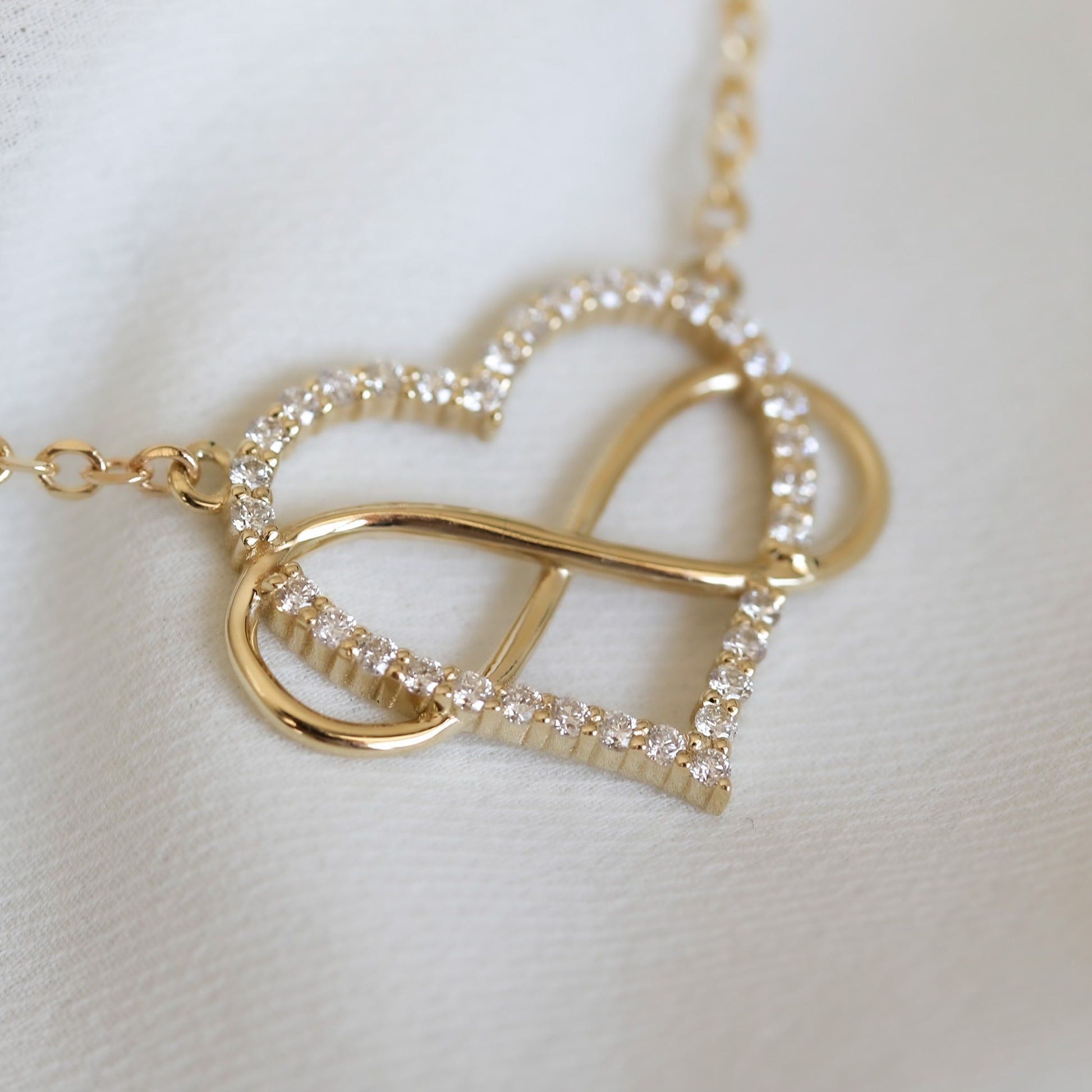 close up of a heart-shaped pendant necklace with a diamonds and gold infinity symbol, attached to a yellow gold chain, displayed on white background