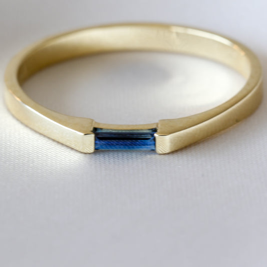 A minimalist style ring with a needle baguette cut blue sapphire set in a 14K yellow gold band, displayed against a white background.