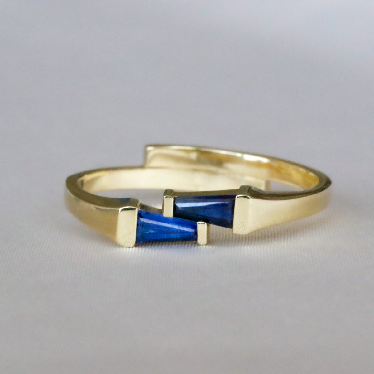 14K yellow gold ring with two tapered baguette blue sapphires set side by side in opposite directions, on a gray background