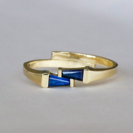 14K yellow gold ring with two tapered baguette blue sapphires set side by side in opposite directions, on a gray background