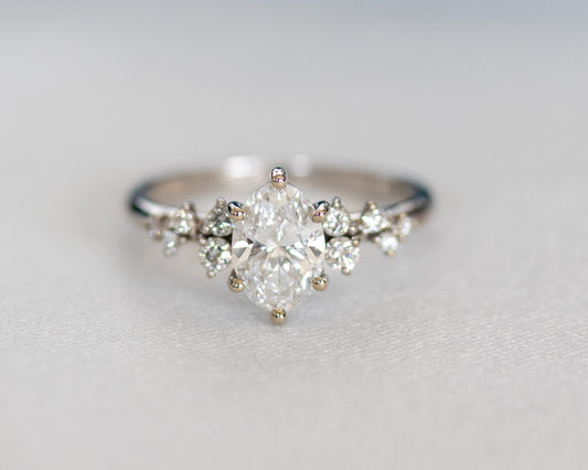 Oval diamond engagement ring with a diamond cluster, in white gold, resting on a soft grey fabric.