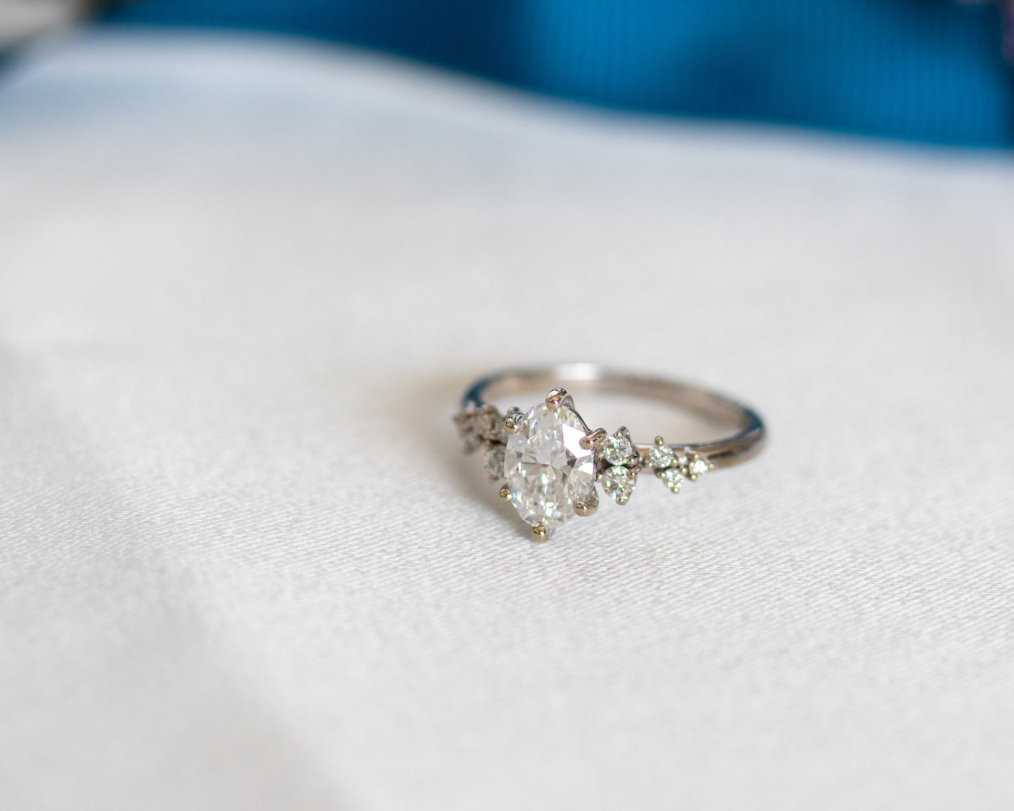 Oval diamond engagement ring with diamond cluster accents resting on a satin fabric.