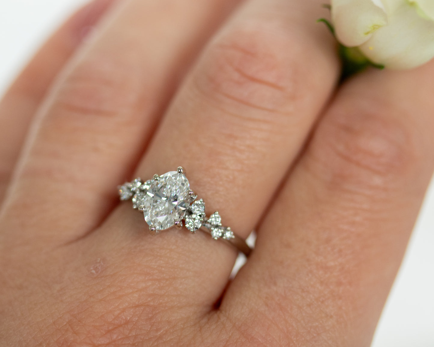 Oval diamond engagement ring worn on a hand, with white flowers in the background.