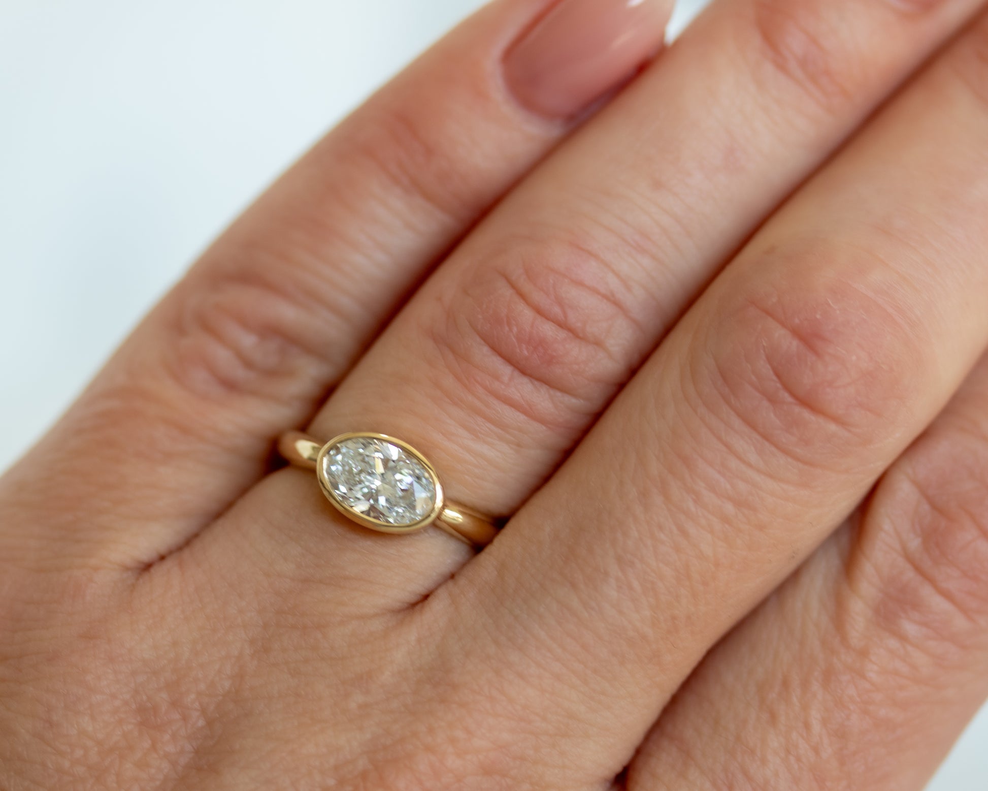 Oval diamond bezel engagement ring stacked with gold bands on hand — modern 14K yellow gold fine jewelry by Rotem Avitan.