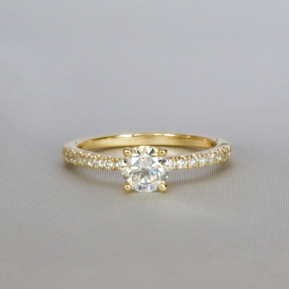 Round brilliant diamond solitaire engagement ring in yellow gold with a pavé diamond band, front view on a gray background