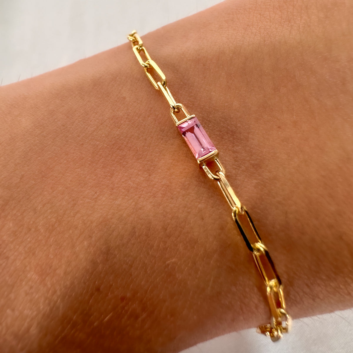 14K yellow gold paperclip chain bracelet with a baguette cut pink sapphire at its center, worn on the wrist