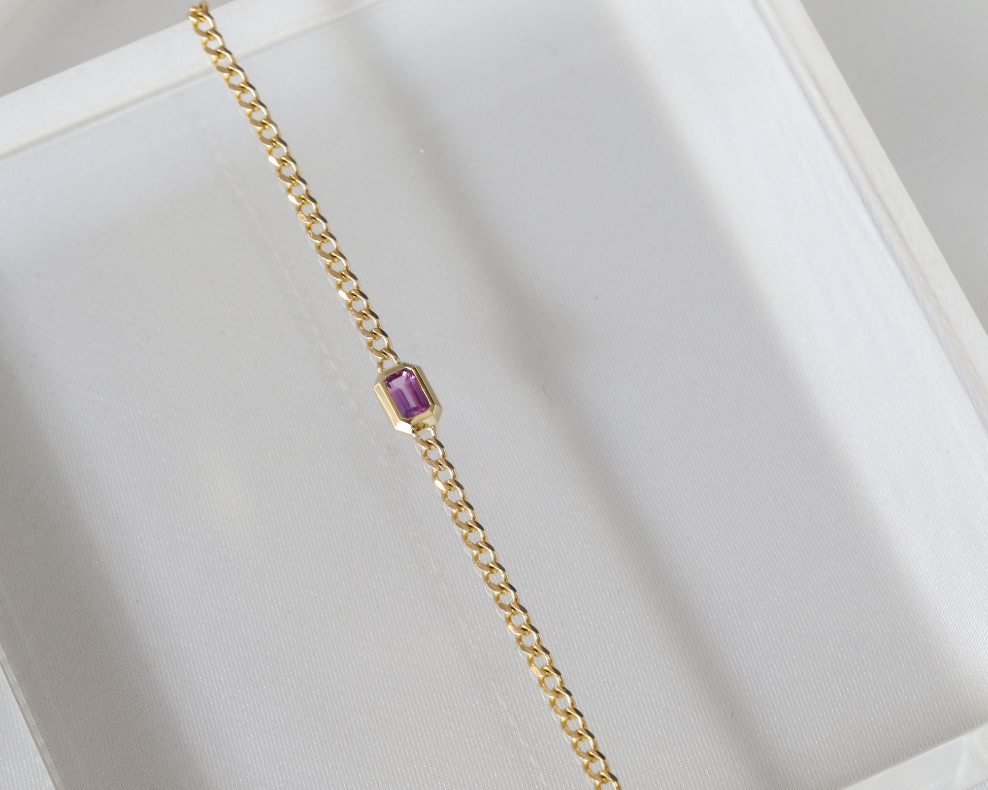 14K yellow gold curb chain bracelet with emerald cut pink sapphire laid flat on a light background