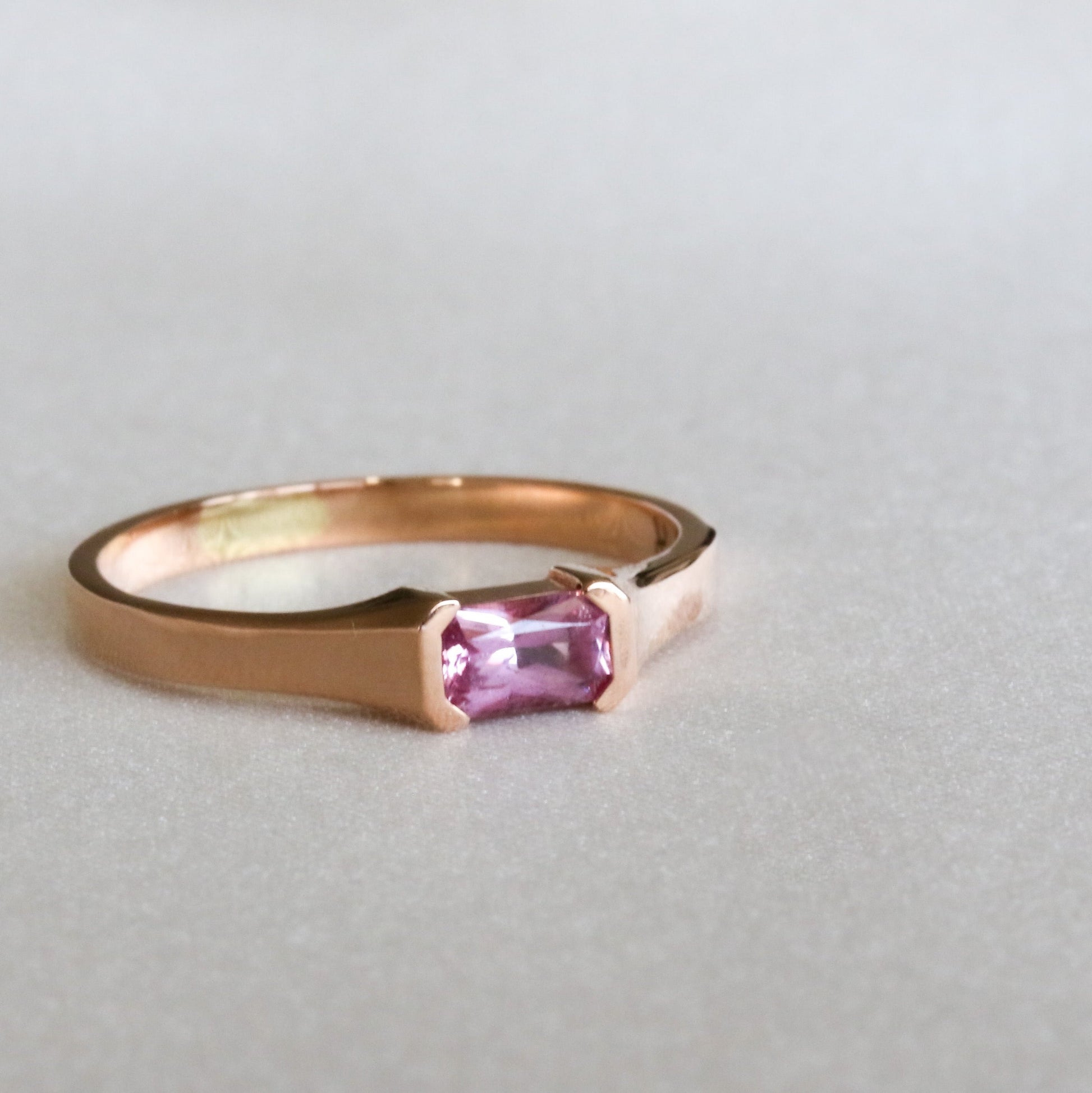 A rose gold ring with a emerald cut natural pink sapphire stone, set in a half bezel setting.