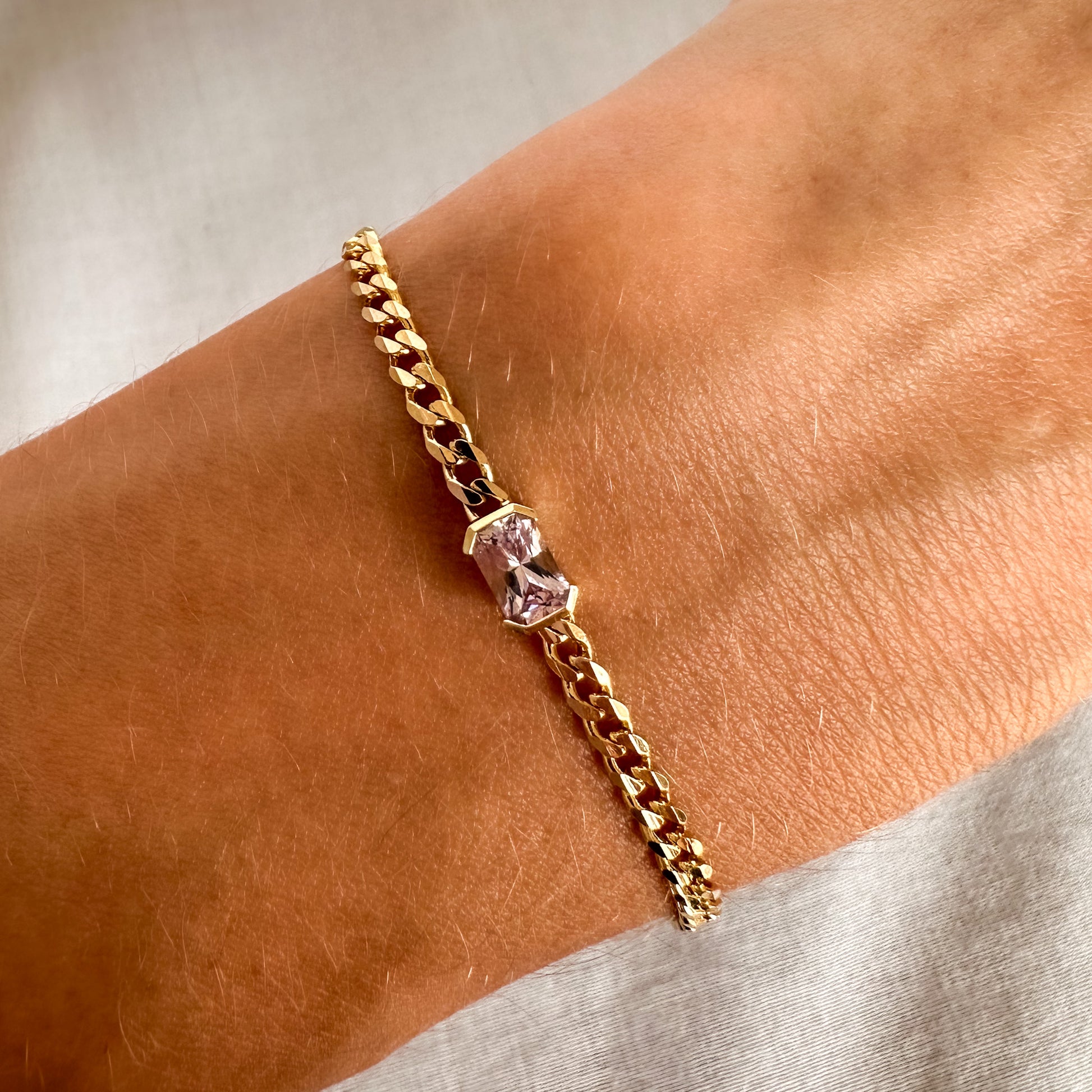 A gold bracelets with a curb chain design one featuring an emerald cut natural pink sapphire displayed on a person's wrist.