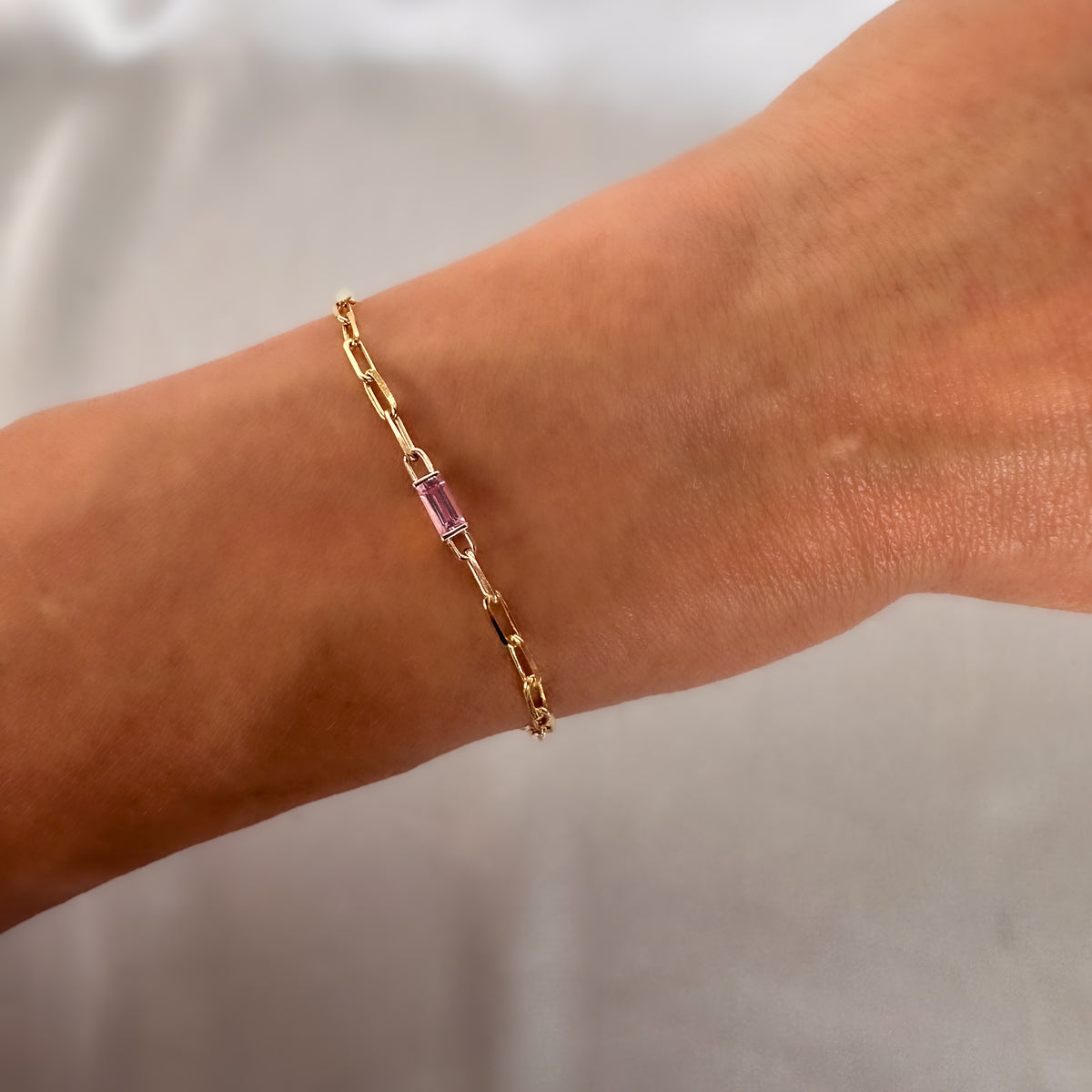 Gold paperclip chain bracelet with a baguette cut pink sapphire, worn on the wrist, showing its size on the hand