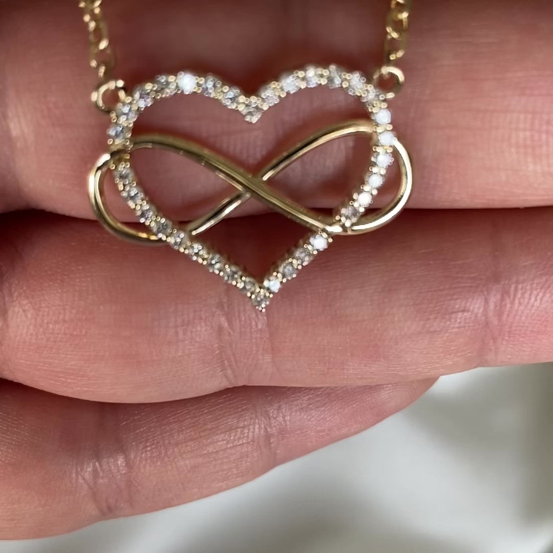 A heart-shaped pendant necklace with a diamonds and gold infinity symbol, attached to a yellow gold chain, displayed in person's hand